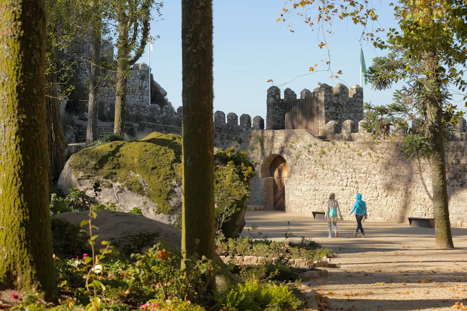 Two people walk toward an ancient stone castle, surrounded by greenery and flowers on a sunny day.