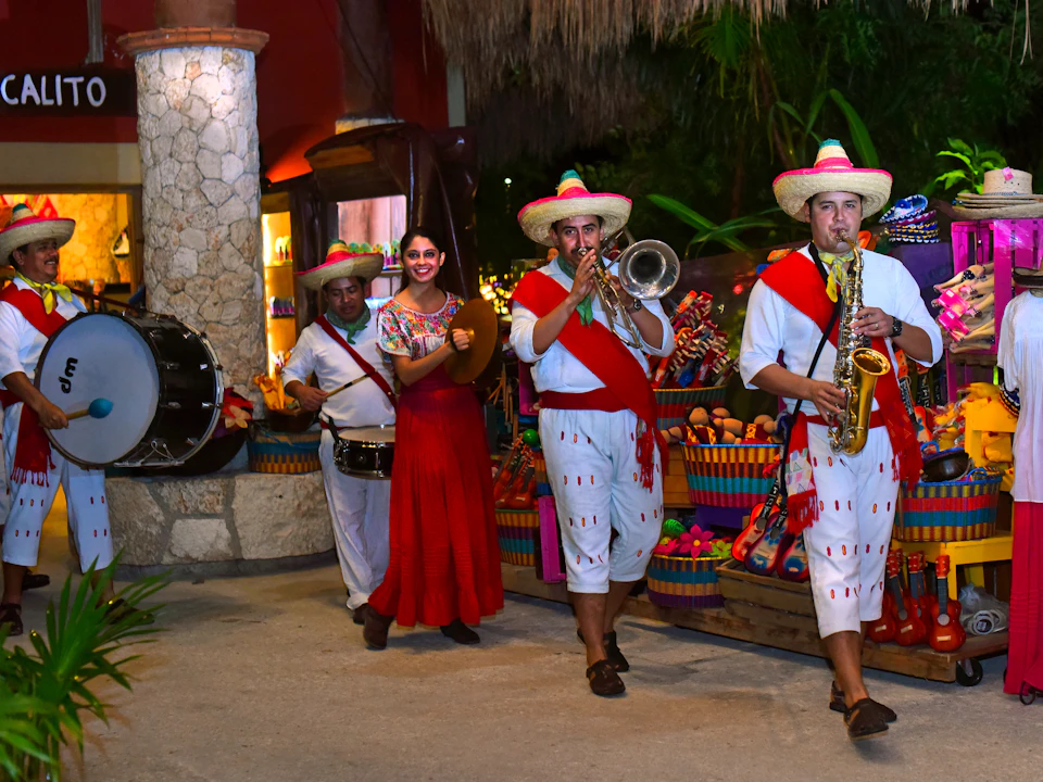 Musicians in traditional attire play instruments while a woman in a red dress smiles, surrounded by colorful market items at night.