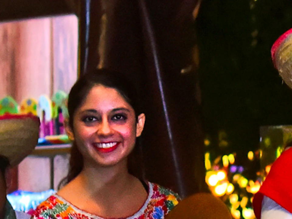 Musicians in traditional attire play instruments while a woman in a red dress smiles, surrounded by colorful market items at night.