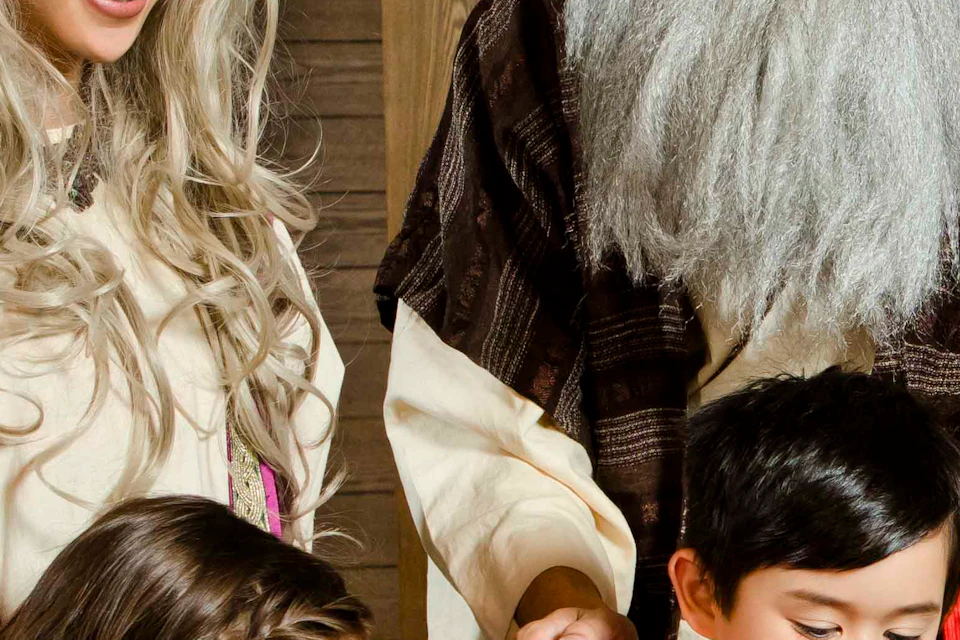 Two adults in medieval costumes with two children use a wooden historical tool; another woman observes, seated at a table.