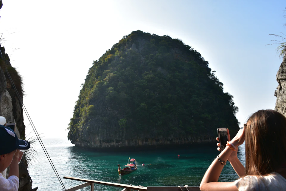 A person holding a phone overlooks a large, tree-covered rock formation rising from the calm sea, with a small boat nearby.
