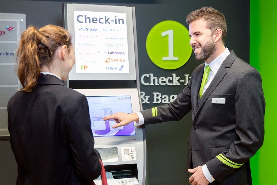 A uniformed airline staff member assists a passenger at a self-check-in kiosk, with a sign reading "Check-In & Bag" behind him.