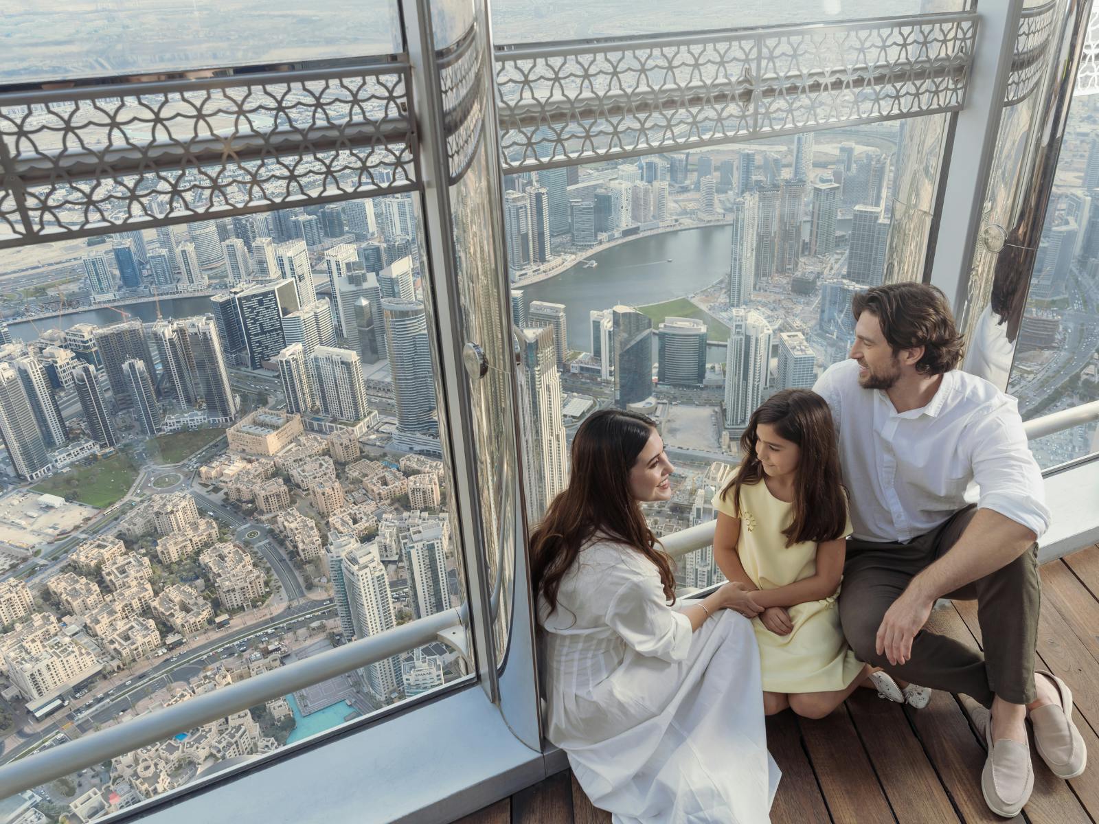 A family of three sits on a wooden floor near large windows, overlooking a cityscape of tall buildings and a waterway below.