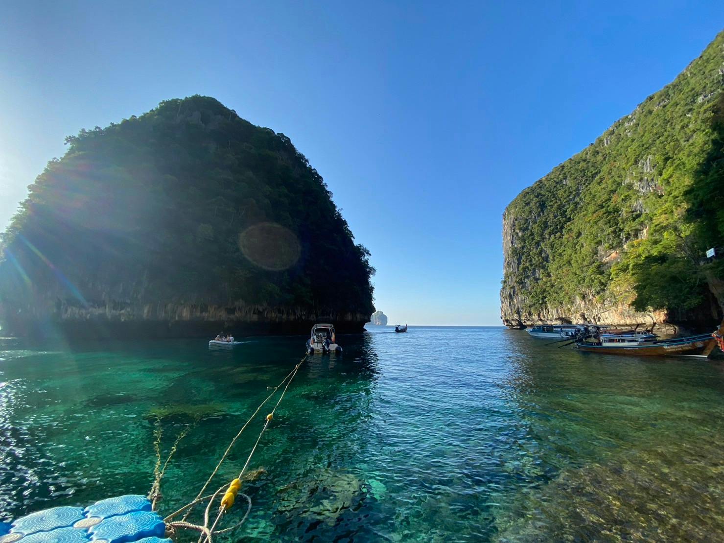 Une baie tranquille avec une eau bleue claire, des bateaux ancrés et des falaises abruptes et verdoyantes de chaque côté, sous un ciel clair.