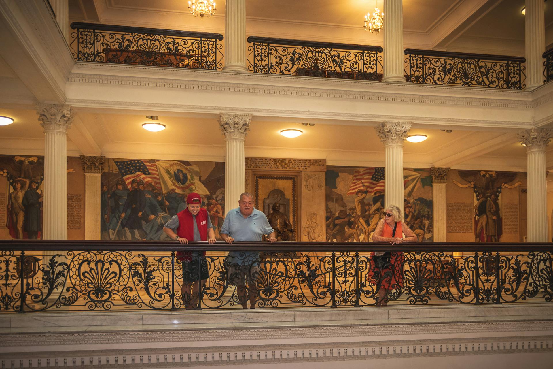 Three people stand on an ornate balcony inside a historic building with murals and flags in the background.