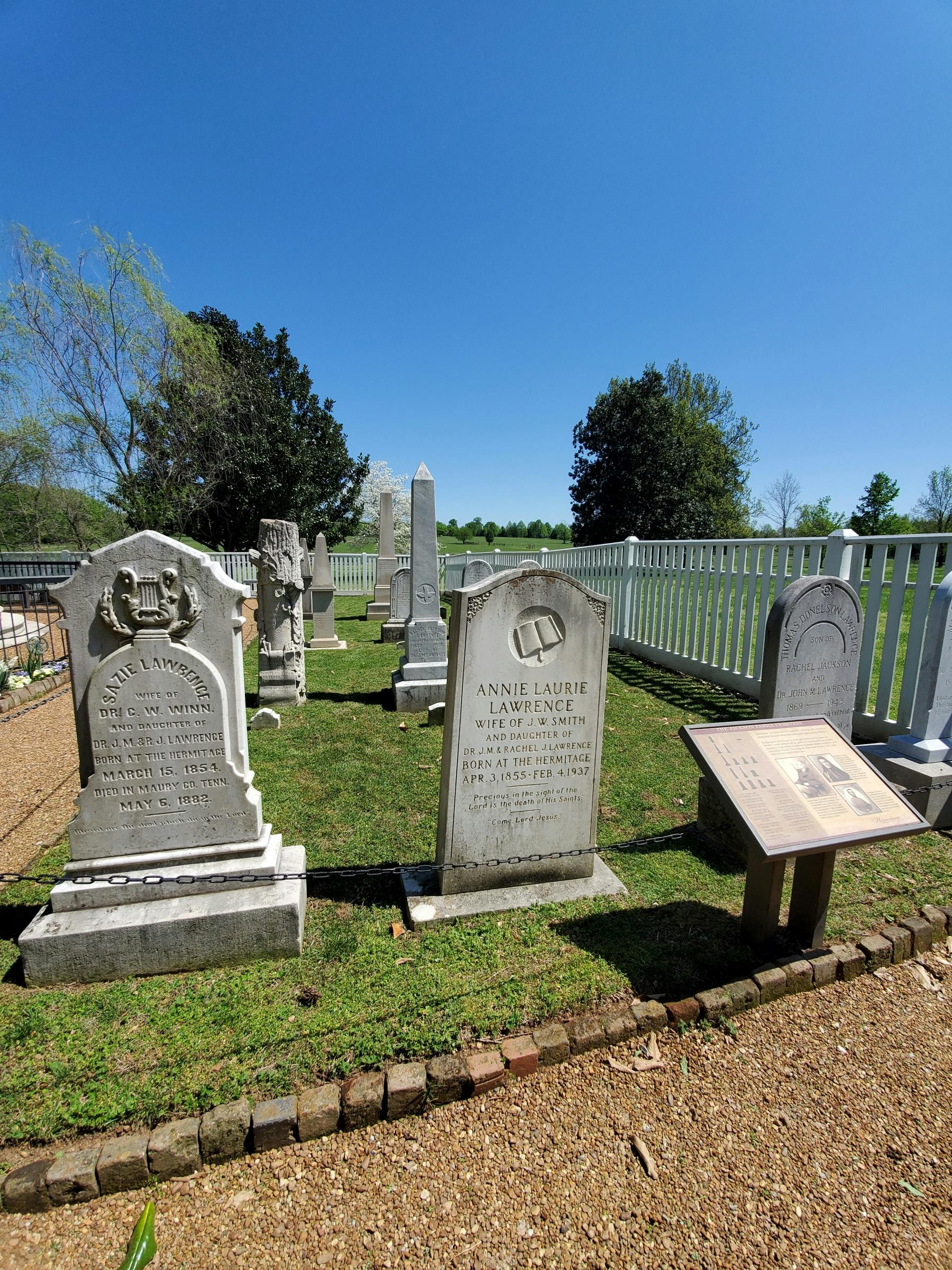 Gravesite with multiple headstones, informational plaque, white fence, and green trees under a clear blue sky.