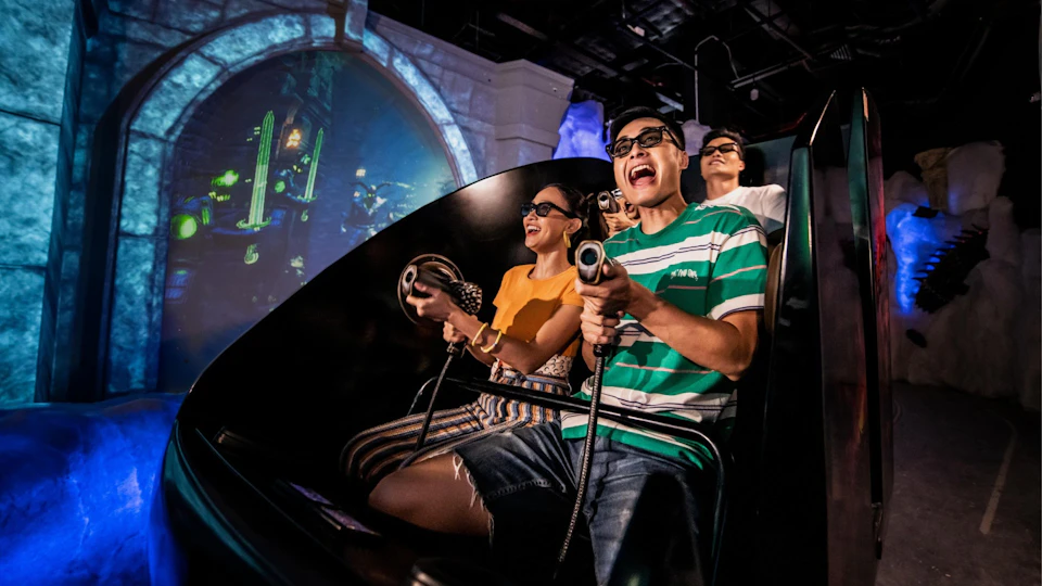 Group of people wearing 3D glasses, smiling and holding laser guns in an indoor amusement ride setting.