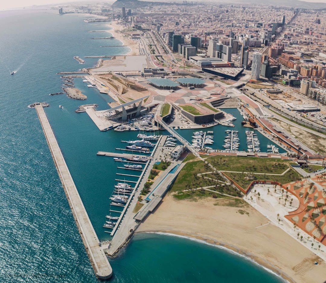 Letecký pohled na pobřežní město s přístavy, loděmi, plážemi a četnými budovami přiléhajícími k velkému nábřeží.