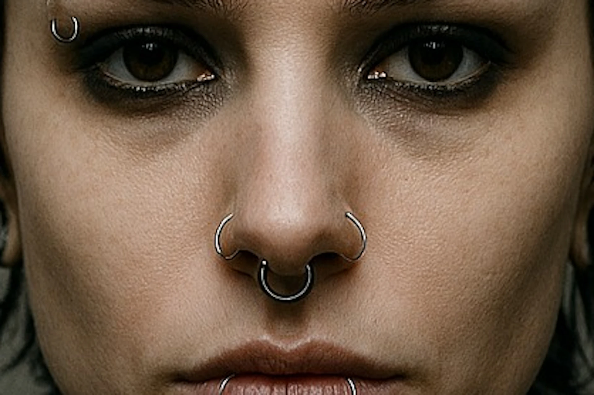 A person with piercings, tattoos, and short dark hair wearing a dark jacket, shown in a neutral background setting.