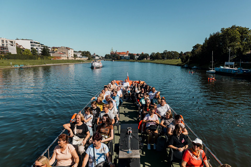 A group of people seated on an open-deck boat traveling along a river on a sunny day, with buildings and trees in the background.