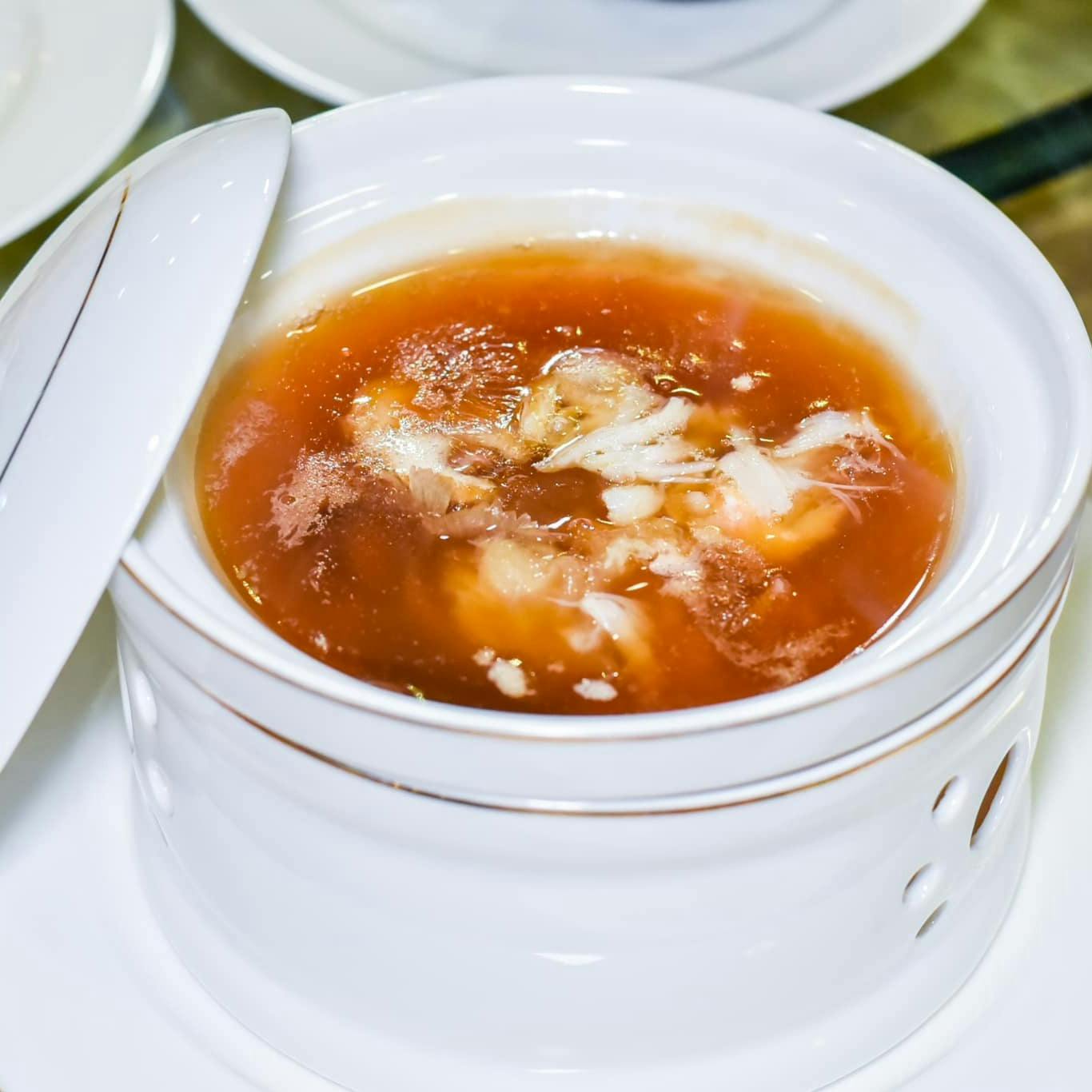 A bowl of soup with a reddish broth and white ingredients, placed in a white ceramic dish on a table.