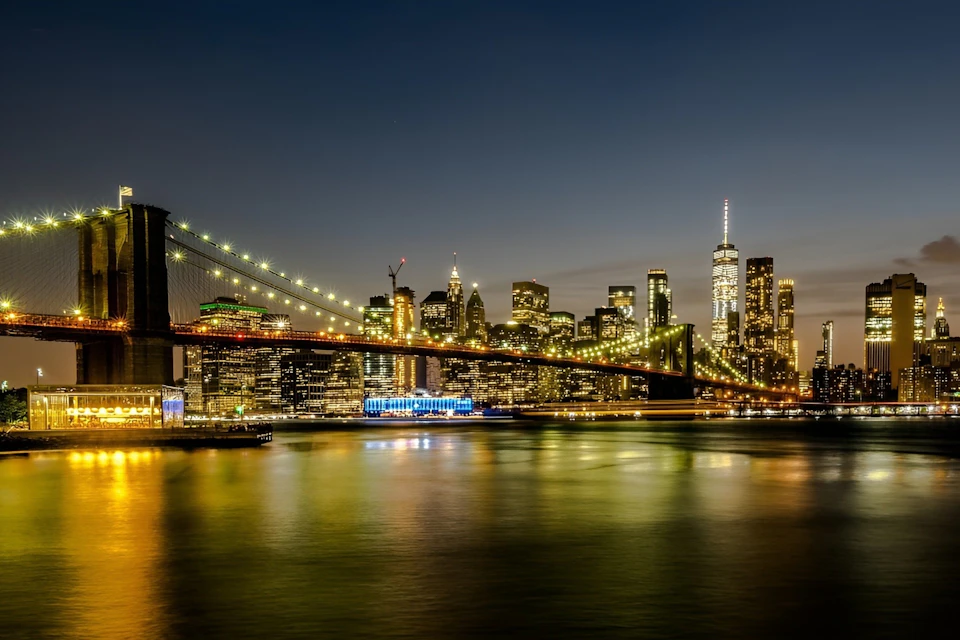 City skyline at night with a lit suspension bridge over a river and illuminated buildings in the background.