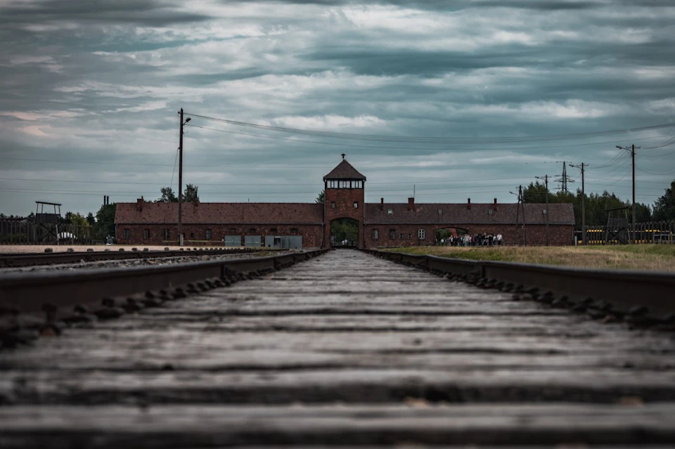 Wooden railroad tracks leading to a brick building with a central entrance tower under a cloudy sky.