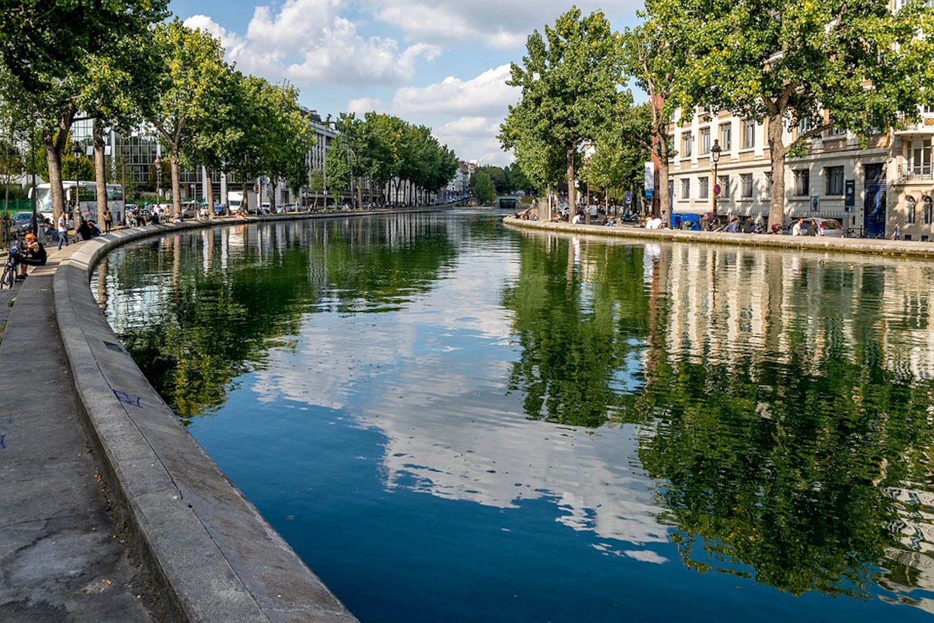 A tree-lined canal with reflective waters, bordered by buildings and a sidewalk, under a partly cloudy sky.