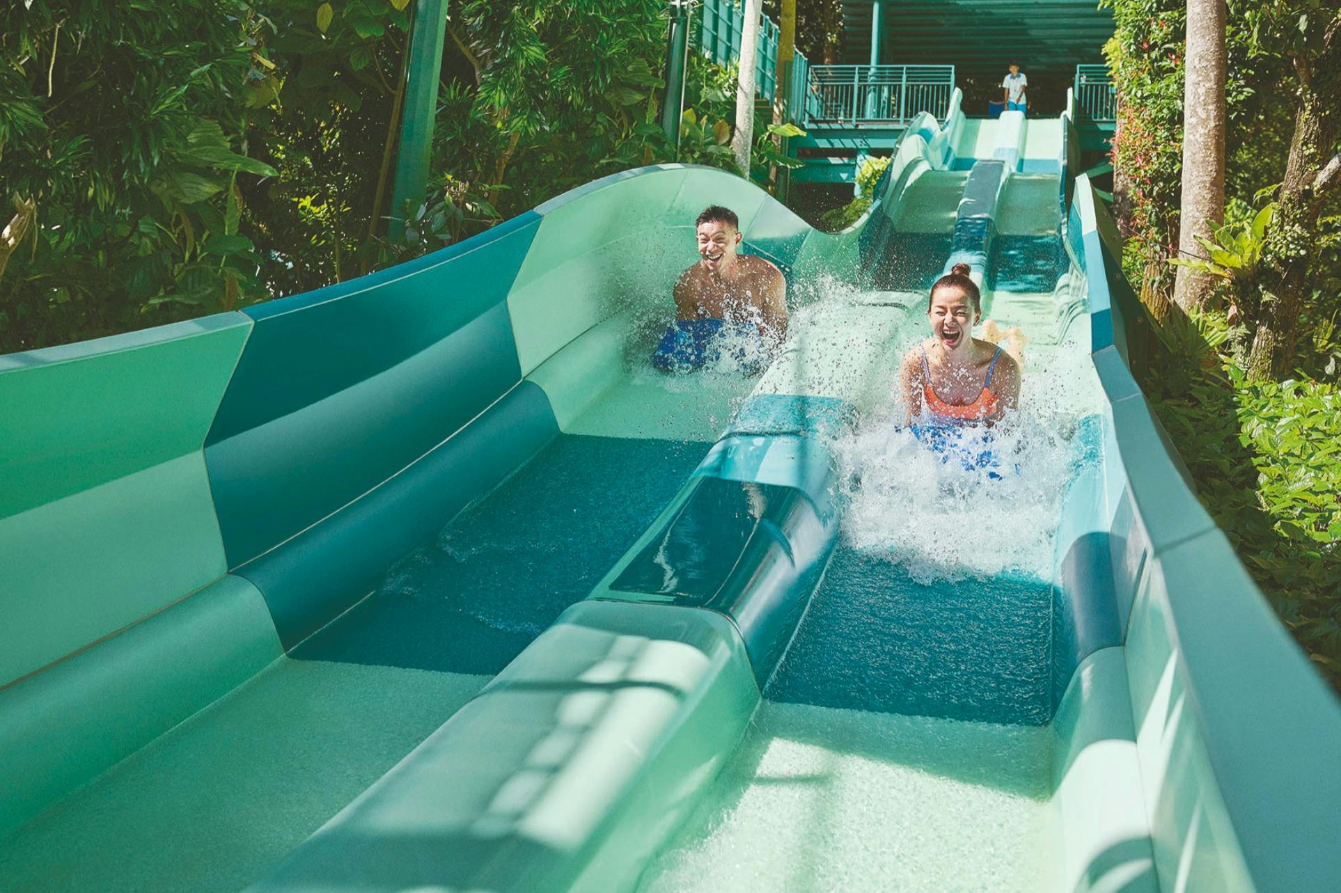 Twee mensen glijden van parallelle glijbanen in een tropische omgeving, lachen en spetteren water.