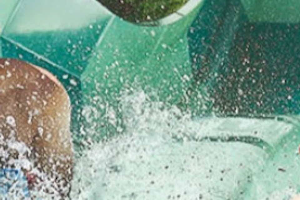 Two people sliding down parallel water slides in a tropical setting, laughing and splashing water.