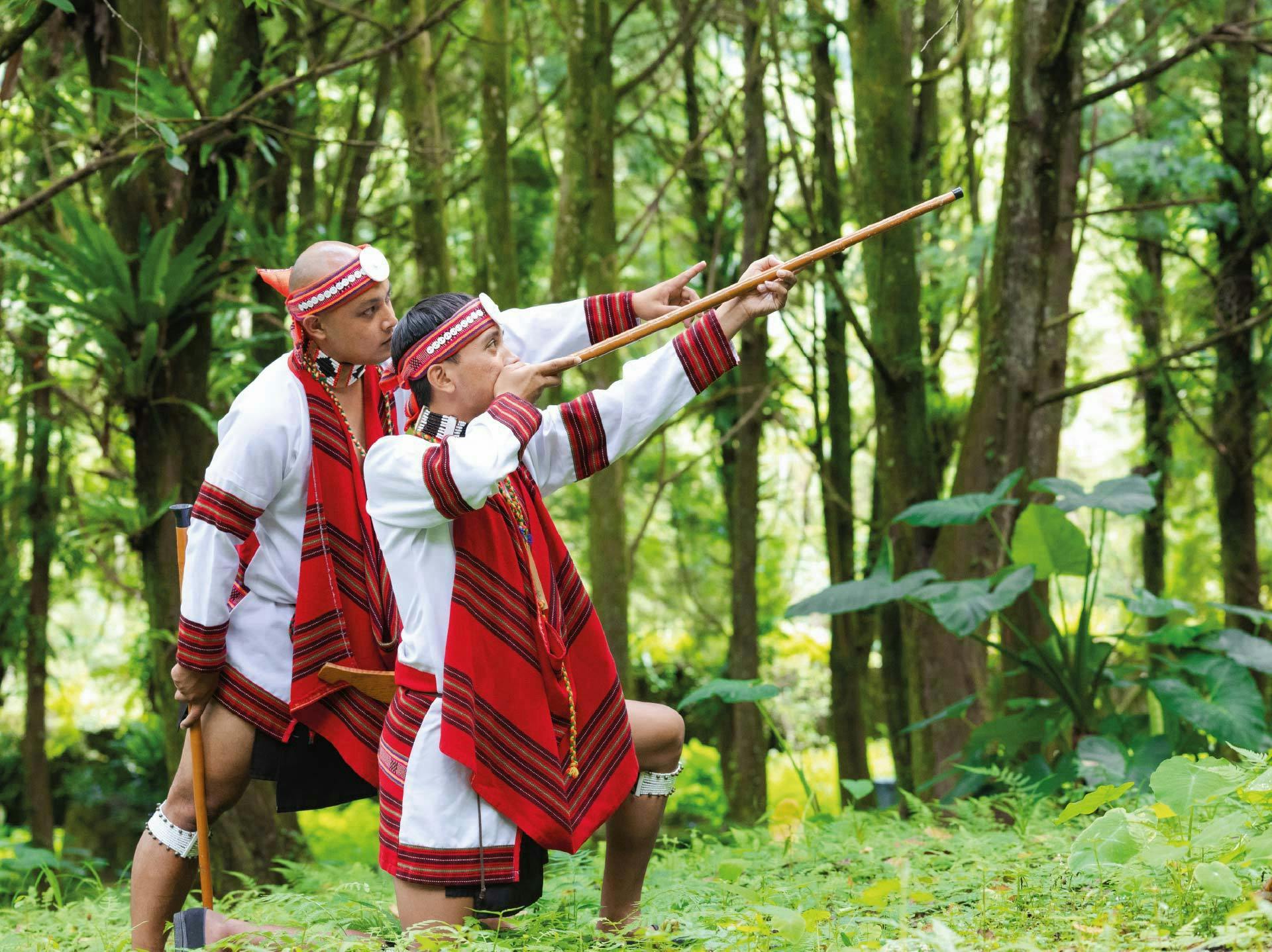 Two individuals in traditional attire with red and white patterns are holding spears in a forest setting.