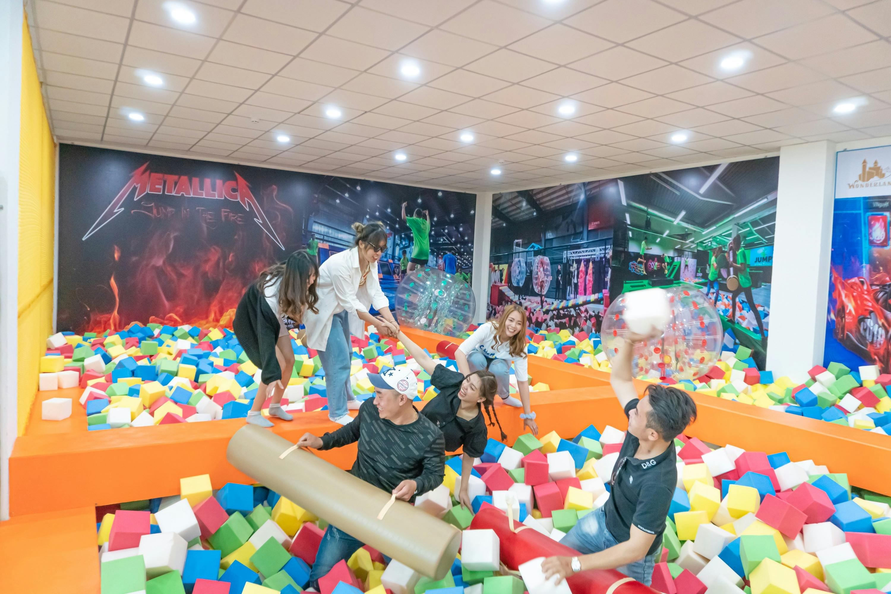 A group of people playing in a colorful foam cube pit indoors, with bright lighting and wall decorations in the background.