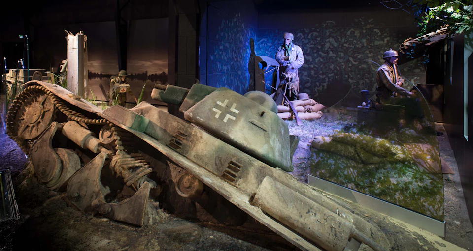 A museum display featuring a damaged military tank and mannequins dressed as soldiers in a World War II trench scene.