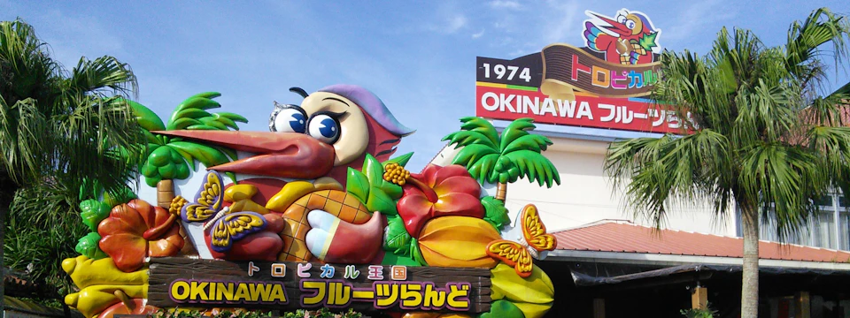 Colorful entrance sign with tropical fruits, a dragon, palm trees, and Japanese text, under a blue sky.