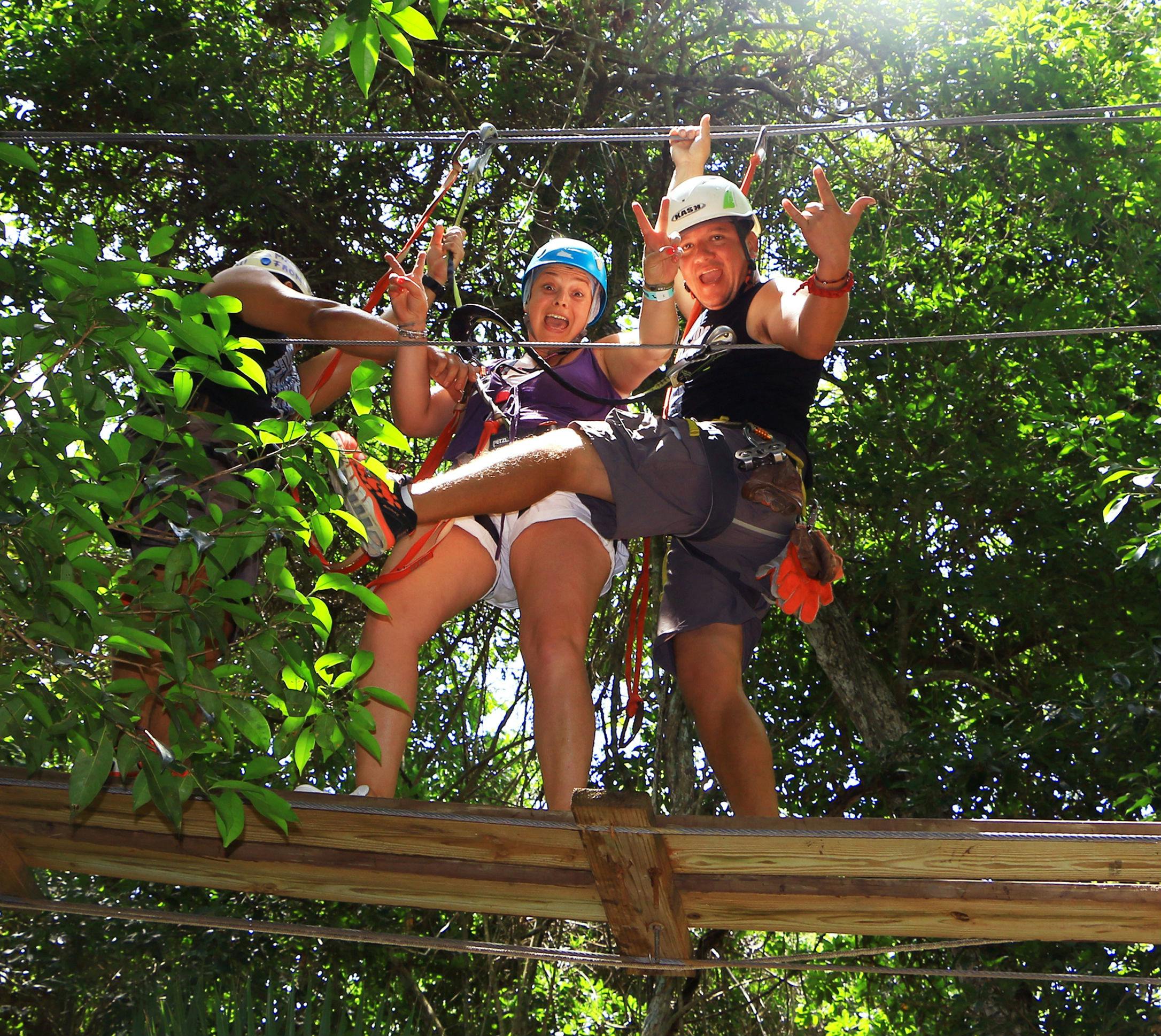 Τρία άτομα με κράνη και ιμάντες στέκονται σε μια ξύλινη πλατφόρμα ανάμεσα στα δέντρα, κάνοντας ενθουσιώδεις χειρονομίες.