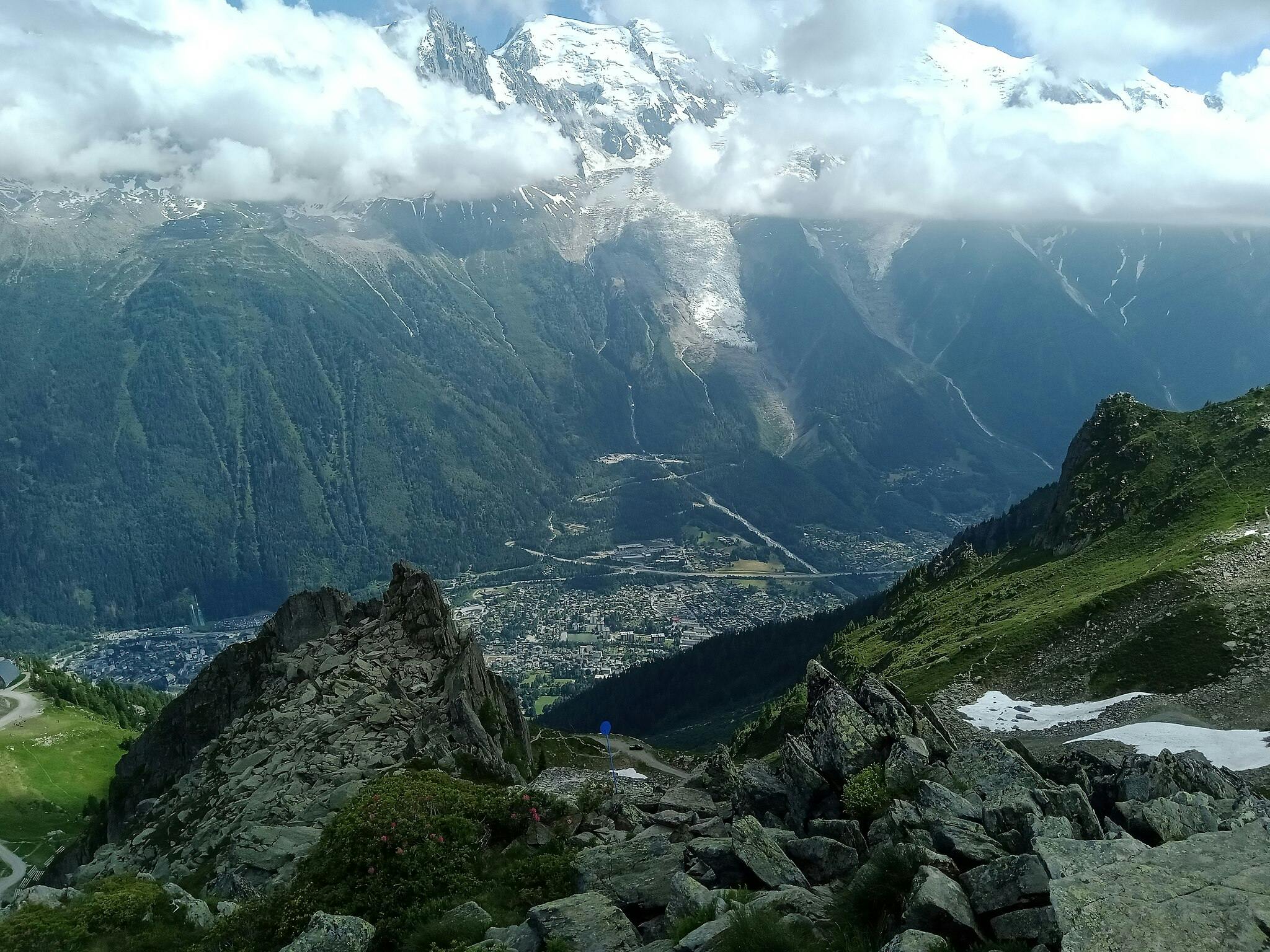 Vue d'une montagne avec un premier plan rocheux, des pentes verdoyantes, une ville en contrebas et des sommets enneigés couverts de nuages au loin.