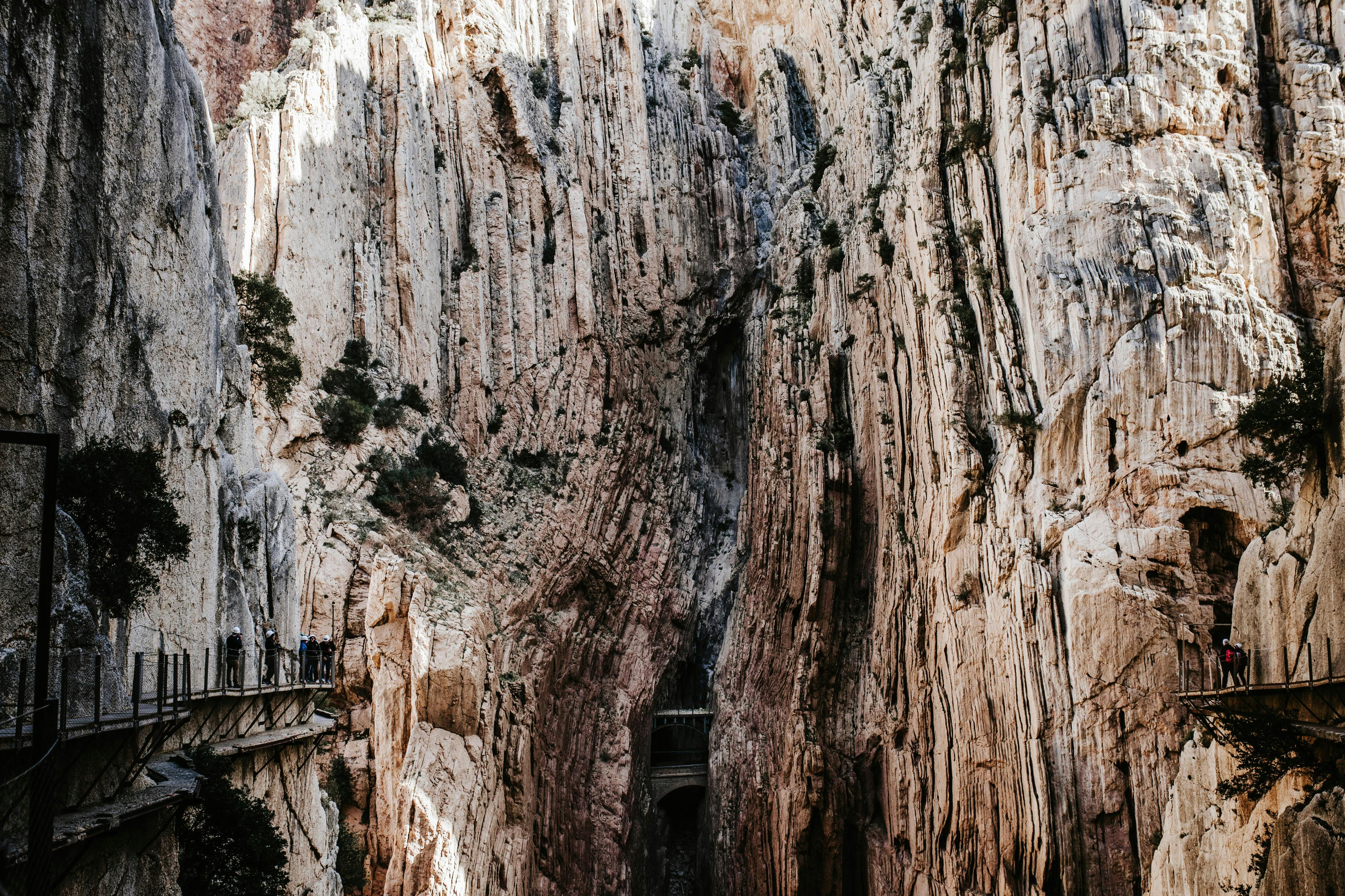 游客站在一条建在锯齿状悬崖边的狭窄人行道上，俯瞰陡峭的岩层和稀疏的植被。
