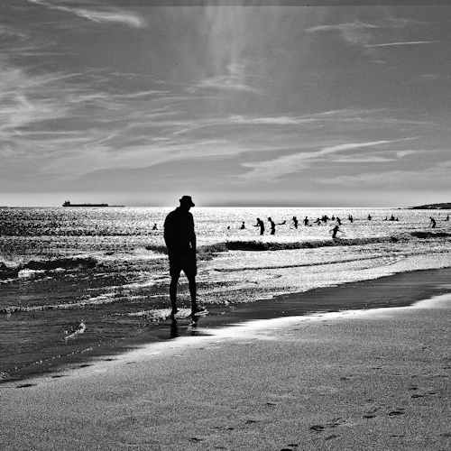 En person går längs en strand med människor som simmar i havet under en ljus, molnig himmel. Fartyg är synliga på avstånd.