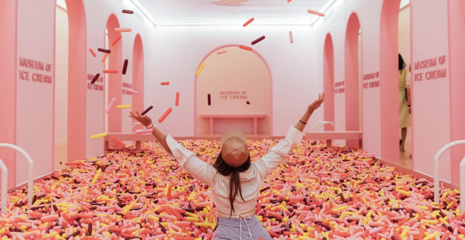 Ένα άτομο με καπέλο ρίχνει πολύχρωμα πλαστικά «πασπαλίσματα» σε ένα ροζ δωμάτιο με την ένδειξη «Μουσείο Παγωτού».