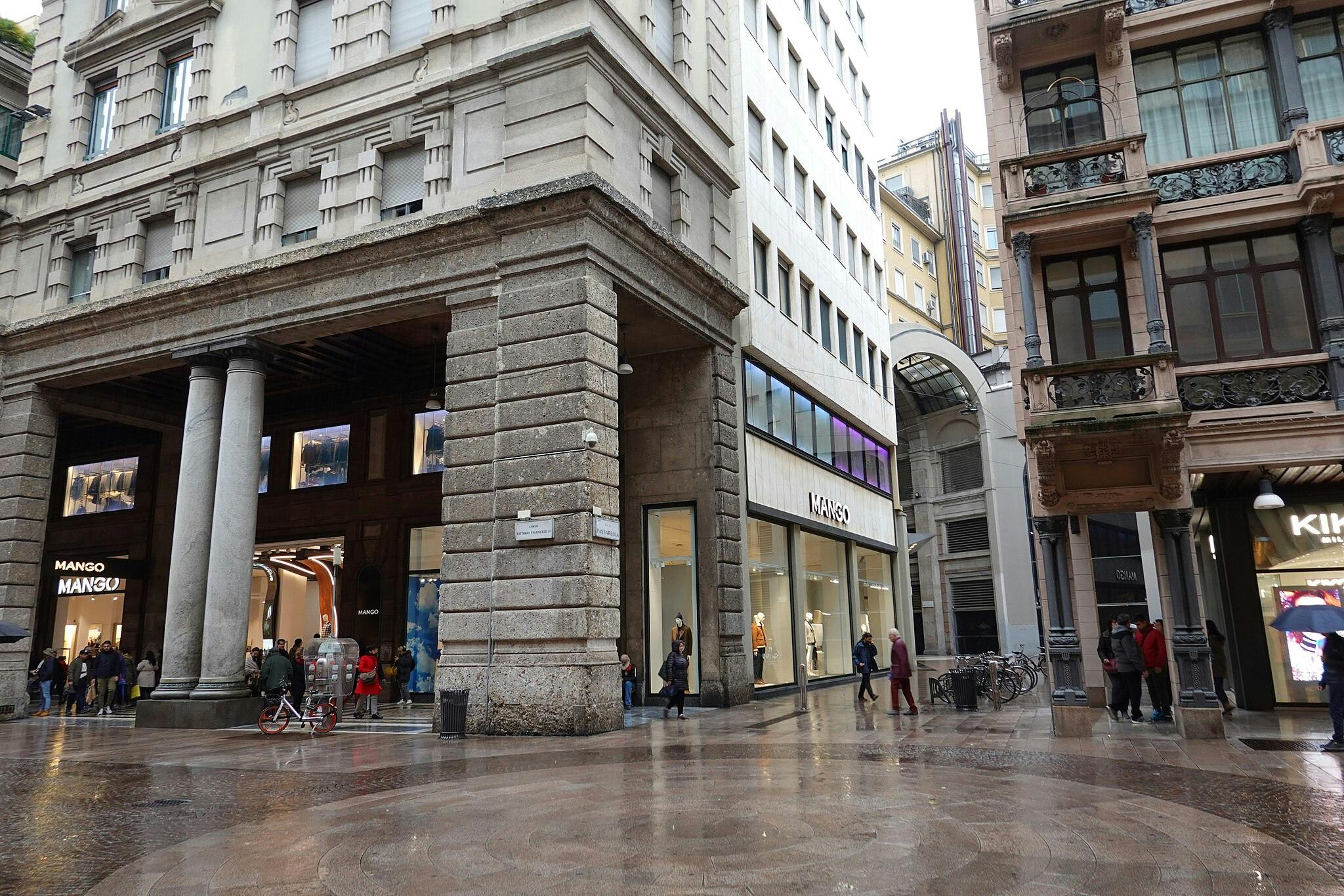 Una scena di strada affollata con persone che camminano vicino all&#39;ingresso di un negozio &#34;Mango&#34;, affiancato da alti edifici in una giornata di pioggia.