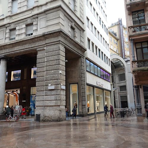 En livlig gatuscen med människor som går nära en "Mango" -butiksentré, flankerad av höga byggnader en regnig dag.