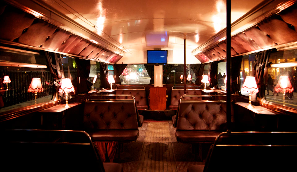 Interior of a vintage bus with tufted leather seating booths, small table lamps, and a TV mounted on the front wall.