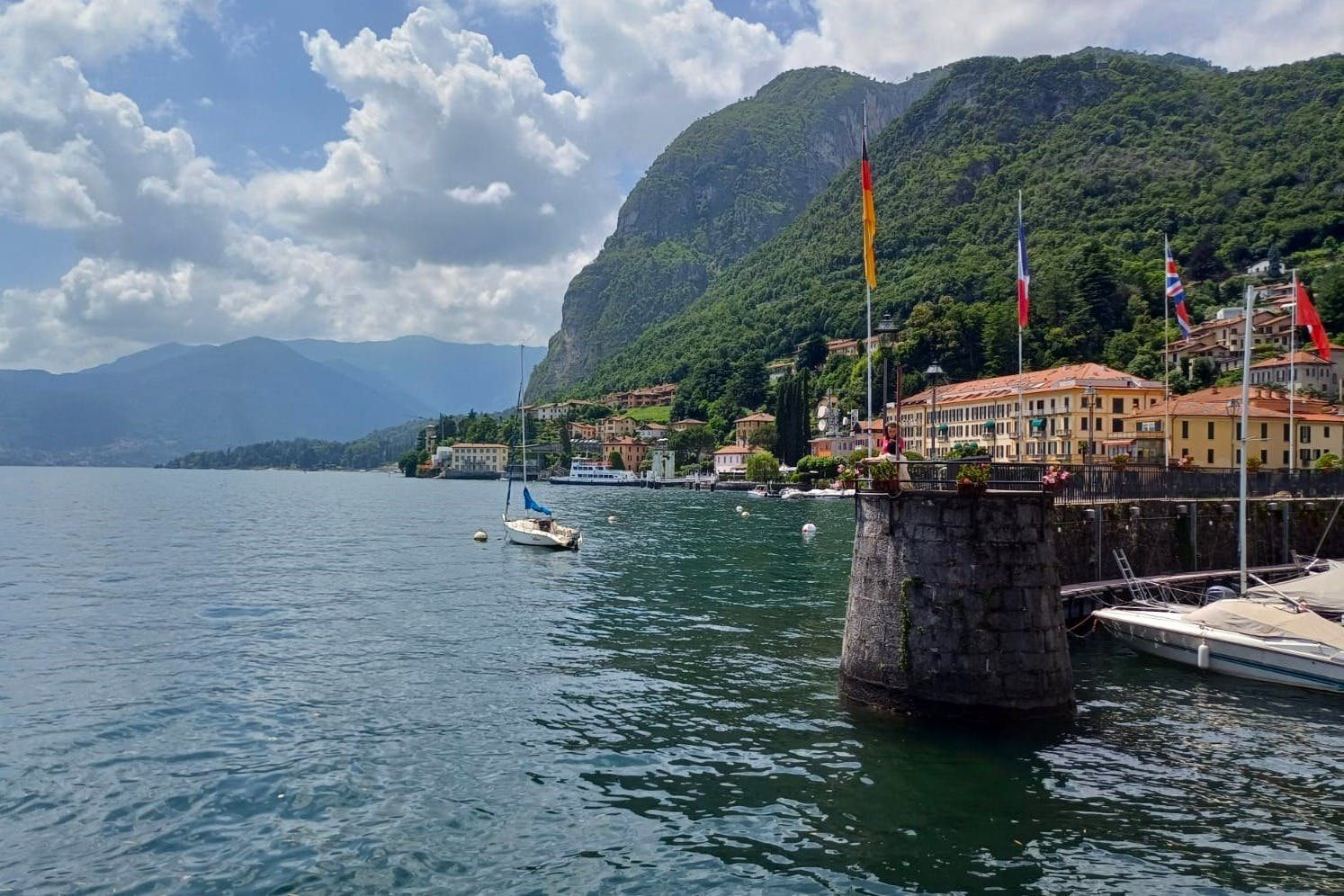 A serene lakeside scene with mountains, a sailboat, waterfront buildings, and flags, under a partly cloudy sky.