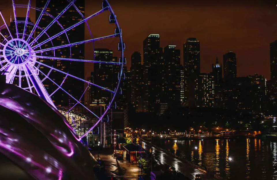 A large Ferris wheel with purple lights next to a waterfront, with illuminated city skyscrapers in the background at night.
