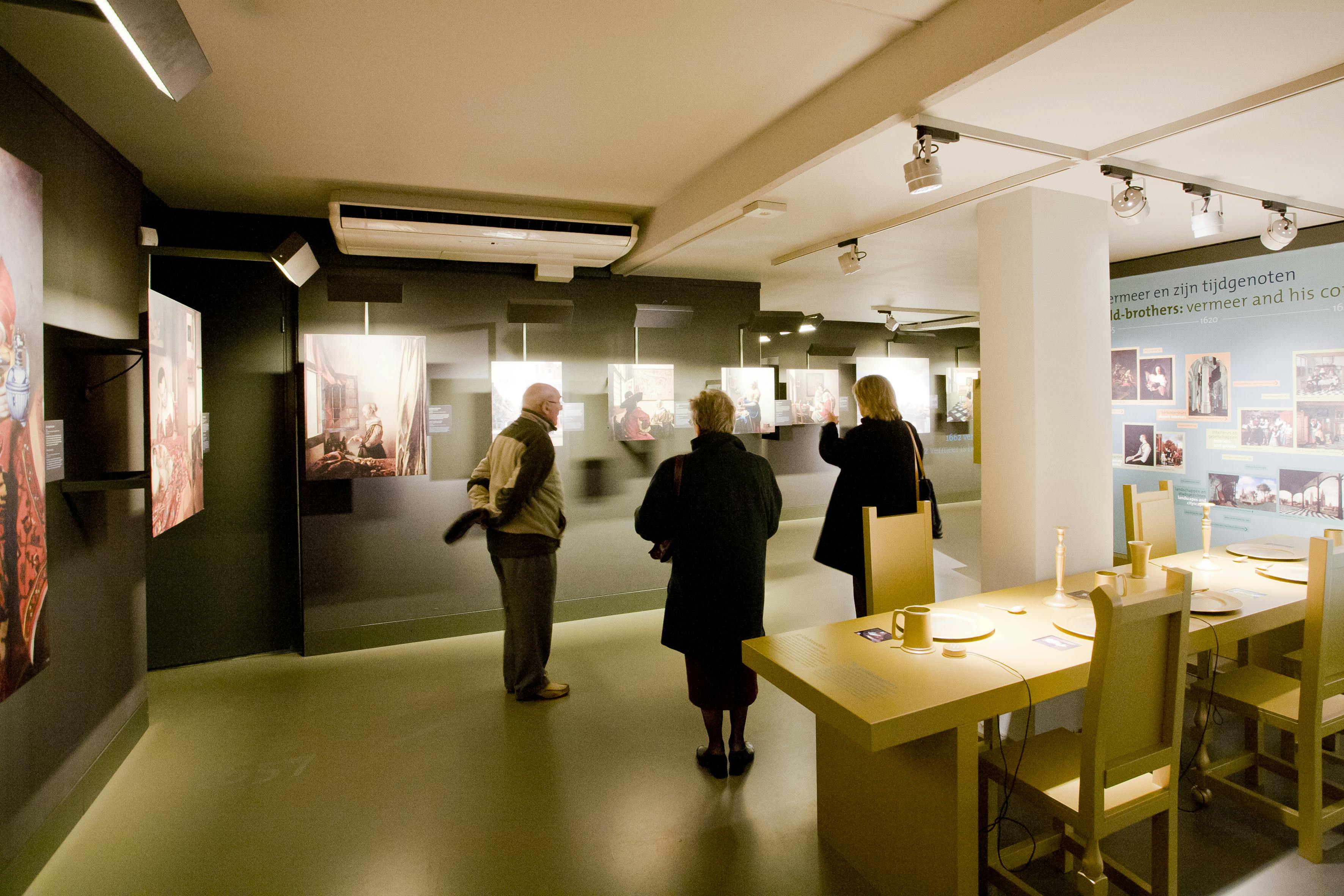 Três pessoas a ver obras de arte emolduradas numa galeria pouco iluminada. Uma mesa com objectos está em primeiro plano, à direita.