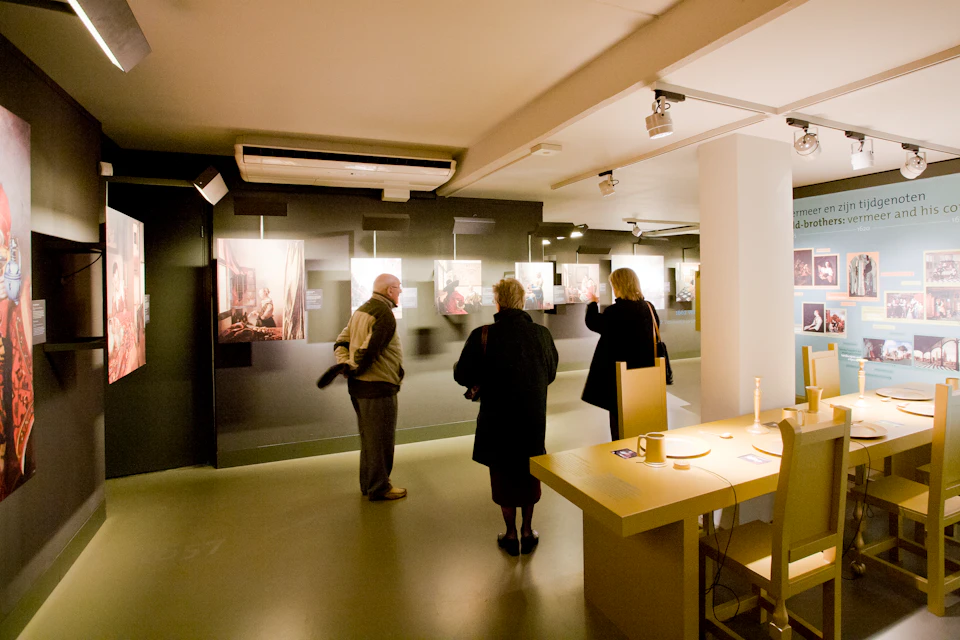 Three people viewing framed artworks in a dimly lit gallery. A table with objects is in the foreground to the right.