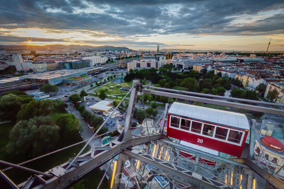 View from a Ferris wheel overlooking a city with green parks, buildings, and a cloudy sky at sunset. Ferris wheel car number 20 is visible.
