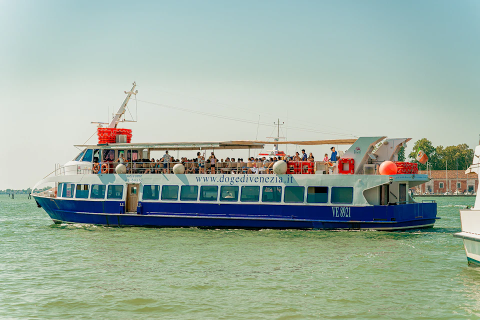 A blue and white passenger boat named VE 821 on the water, with people on board and the website "dogedivenezia.it" displayed.