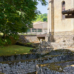 Forntida stenruiner med mossa, bredvid ett stort träd och en gammal byggnad. I bakgrunden finns ett staket och moderna byggnader.