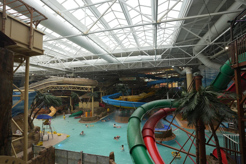 Indoor water park with a pool, waterslides, palm trees, and people swimming under a high ceiling with metal beams.