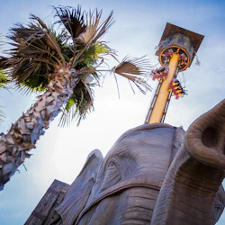 Lågvinkelvy av en palm, en stor elefantskulptur och ett torn med människor. Blå himmel i bakgrunden.