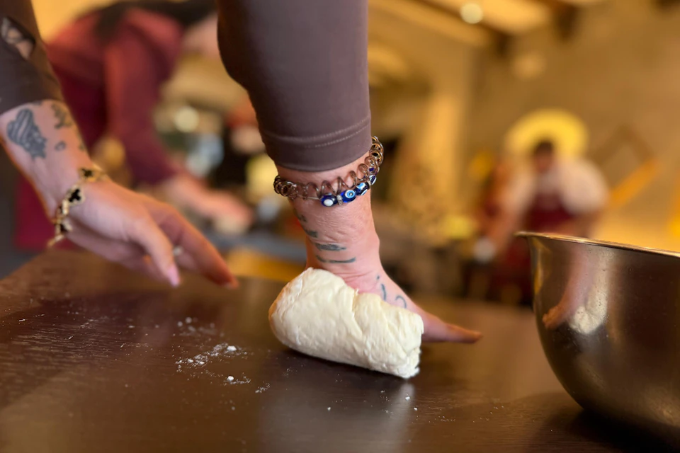 A person kneading dough on a dark surface, with a bracelet visible on their wrist, in a blurred kitchen environment.