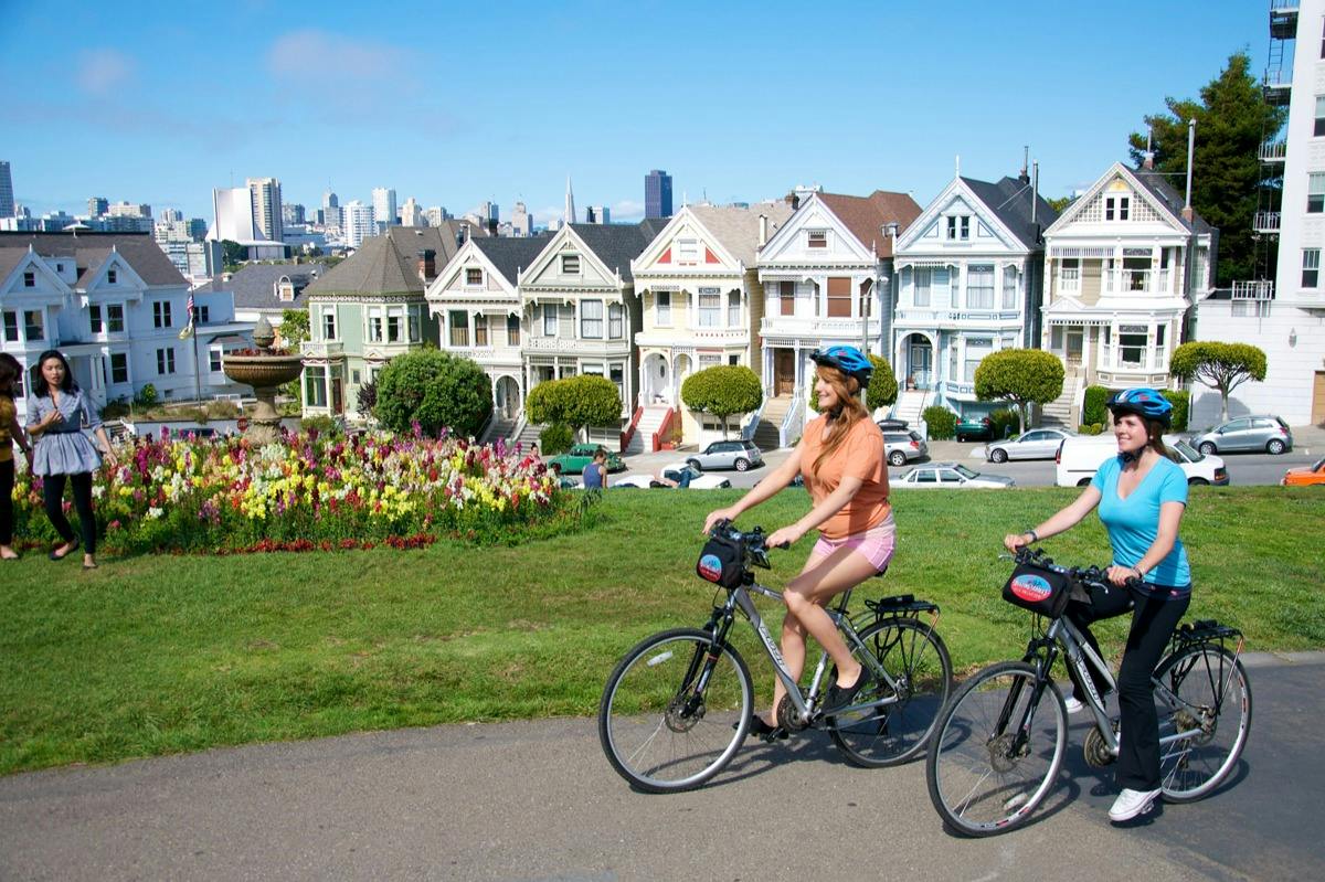 Two people riding bicycles in a park with colorful houses in the background under a clear blue sky.