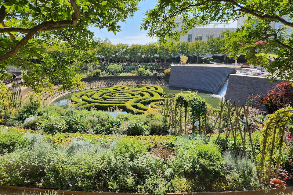 A formal garden with intricate hedges forming a spiral maze, surrounded by lush greenery and a waterfall in the background.