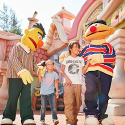 Två barn ler och håller hand med utklädda karaktärer framför en nyckfull byggnad.