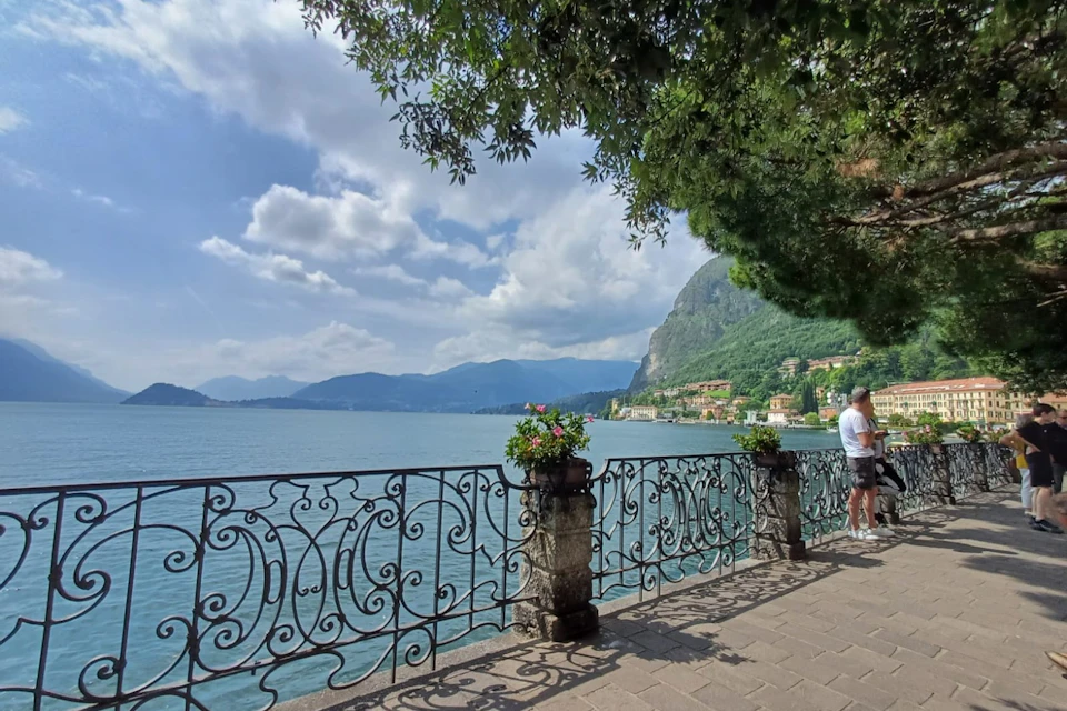 A man is standing by an ornate railing overlooking a lake, with mountains in the distance, under a partly cloudy sky.