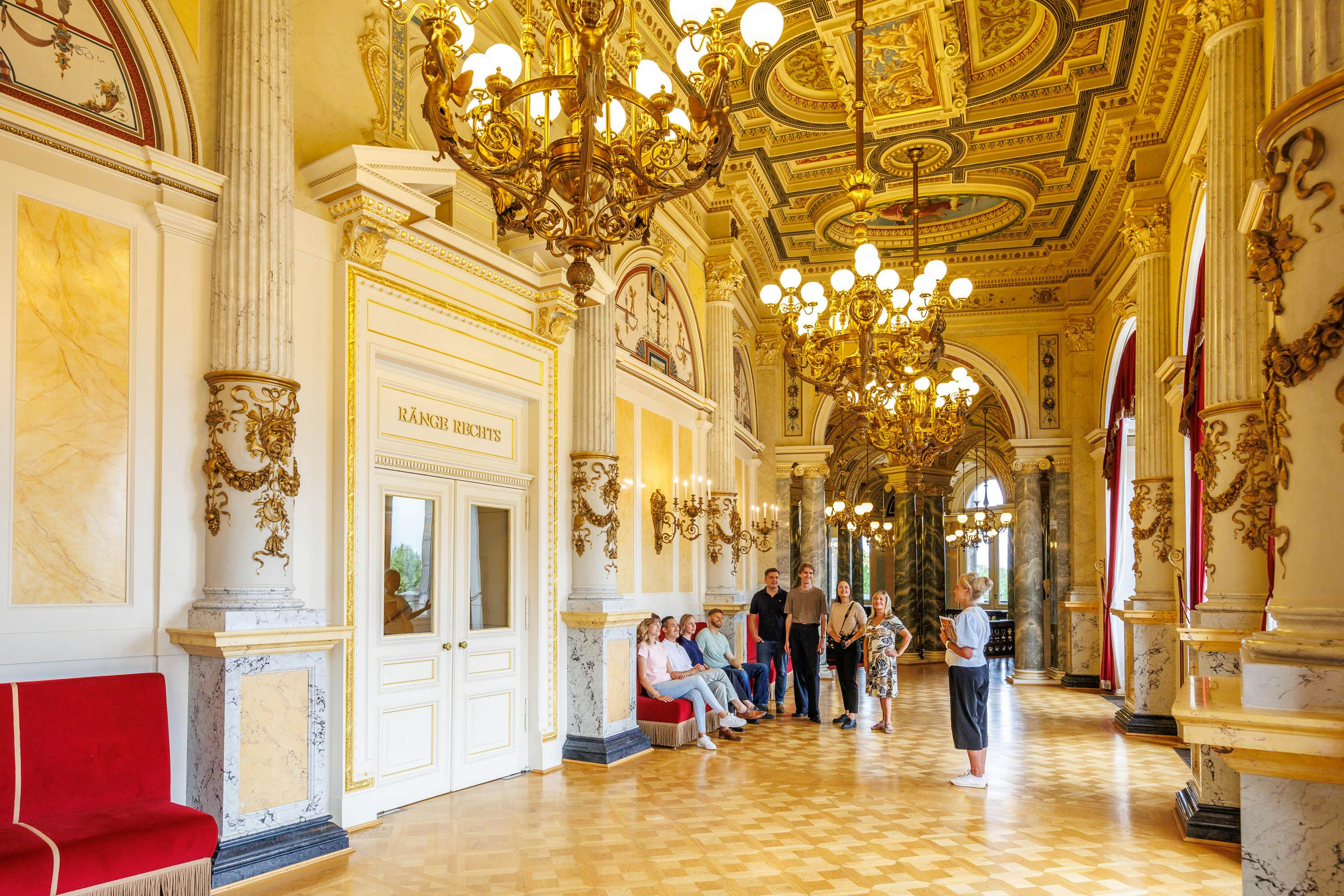 Um grupo de pessoas num salão ornamentado com lustres, decorações douradas e uma placa com a inscrição «Kaiser Box» numa porta.