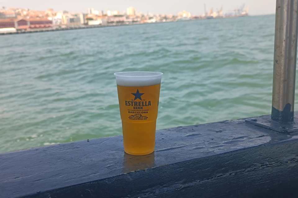 A cup of Estrella Damm beer sits on a ledge with a body of water and a cityscape in the background.