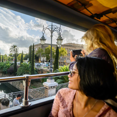 Zwei Personen in einem offenen Bus, von denen die eine einen malerischen Park mit hohen Bäumen, einer Brücke und Gebäuden unter einem bewölkten Himmel fotografiert.