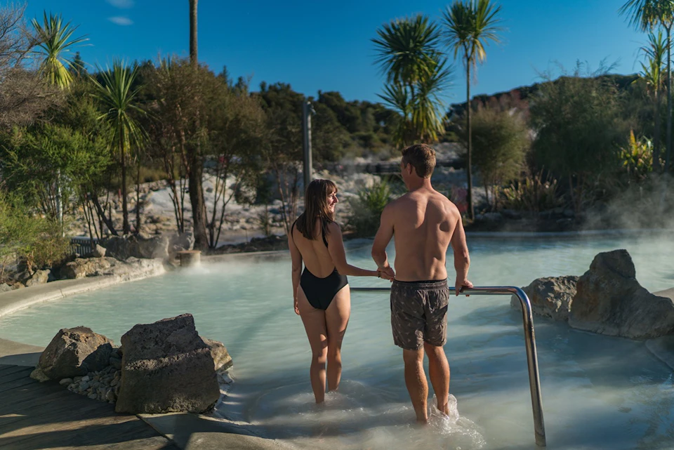 Two people in swimwear standing in a natural geothermal pool surrounded by rocks and trees under a clear sky.